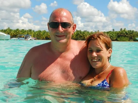 Manuela y Rainer, sonriendo en el agua turquesa con paisaje tropical al fondo.