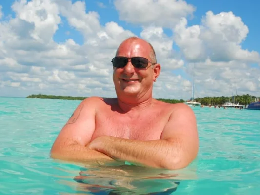 Rainer, sonriendo en el agua con gafas de sol y paisaje caribeño detrás.
