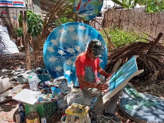 Artista pintando en un taller al aire libre rodeado de pinturas marinas.